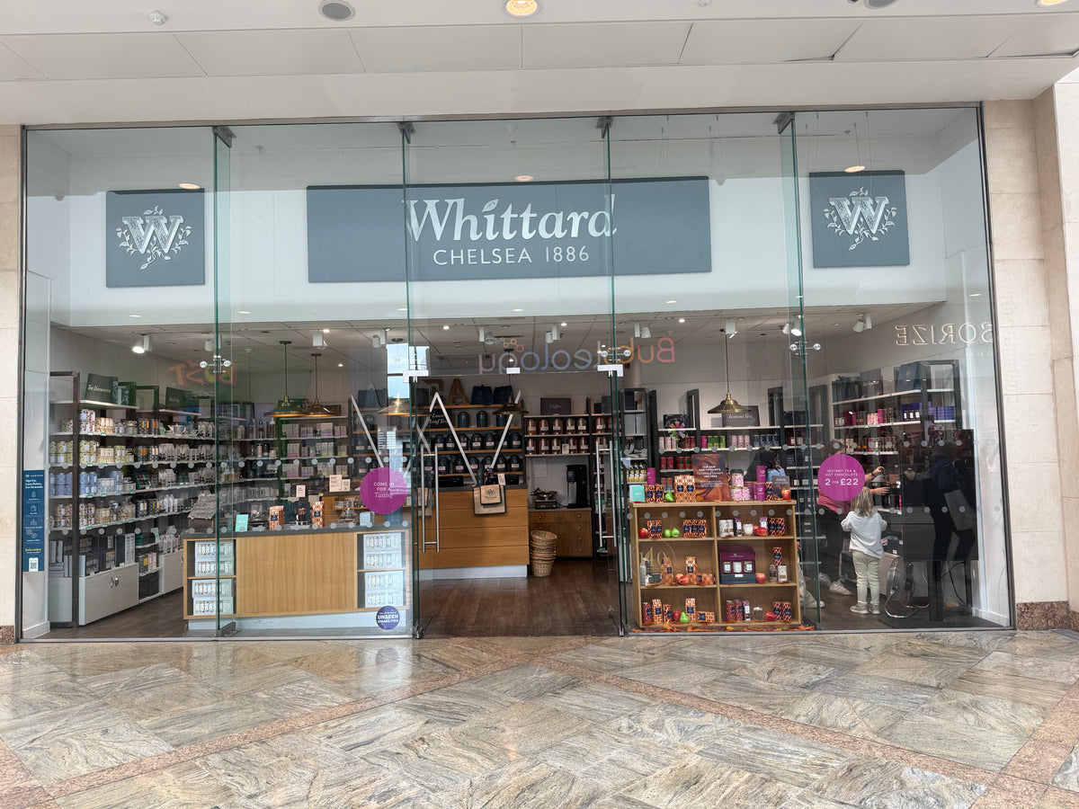 Glass-fronted Whittard Chelsea 1886 shop in a mall, displaying tea, coffee, and gift products on shelves, with a counter and shoppers visible inside.