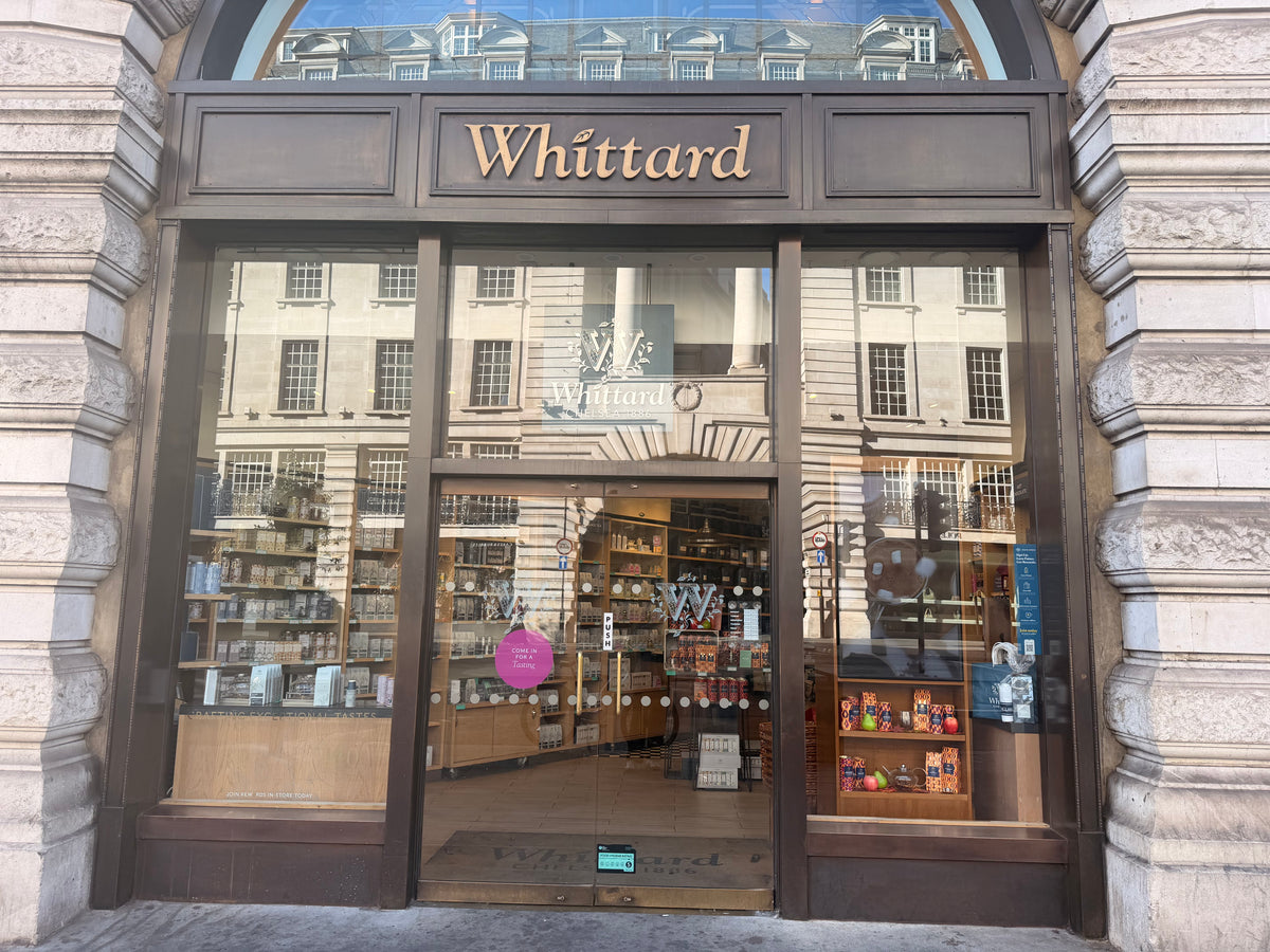 Storefront of Whittard, a tea and coffee shop, with large glass windows displaying shelves of products. The reflection of a building is visible in the glass. The shop is located in a stone building with an arched entrance.