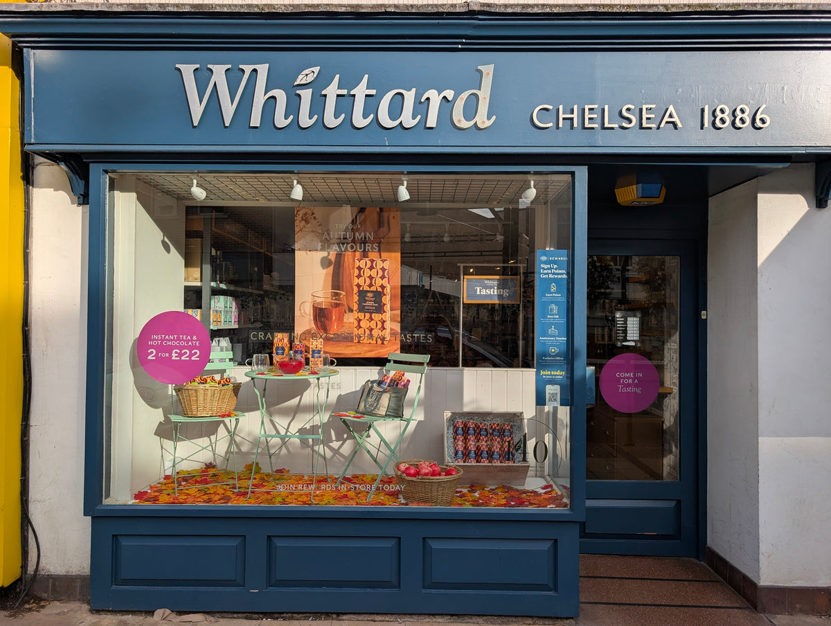 Shopfront of Whittard Chelsea 1886 with a large window display showing tea products, a table with two chairs, baskets, and colorful items. Promotional signs and inviting decor are visible.