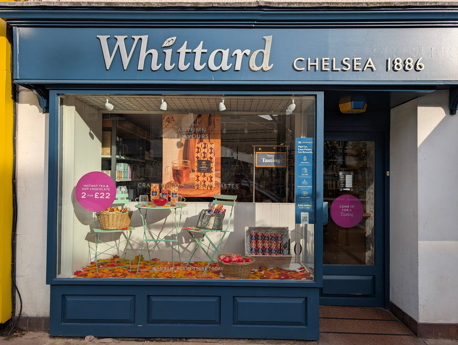 Shopfront of Whittard Chelsea 1886 with a large window display showing tea products, a table with two chairs, baskets, and colorful items. Promotional signs and inviting decor are visible.