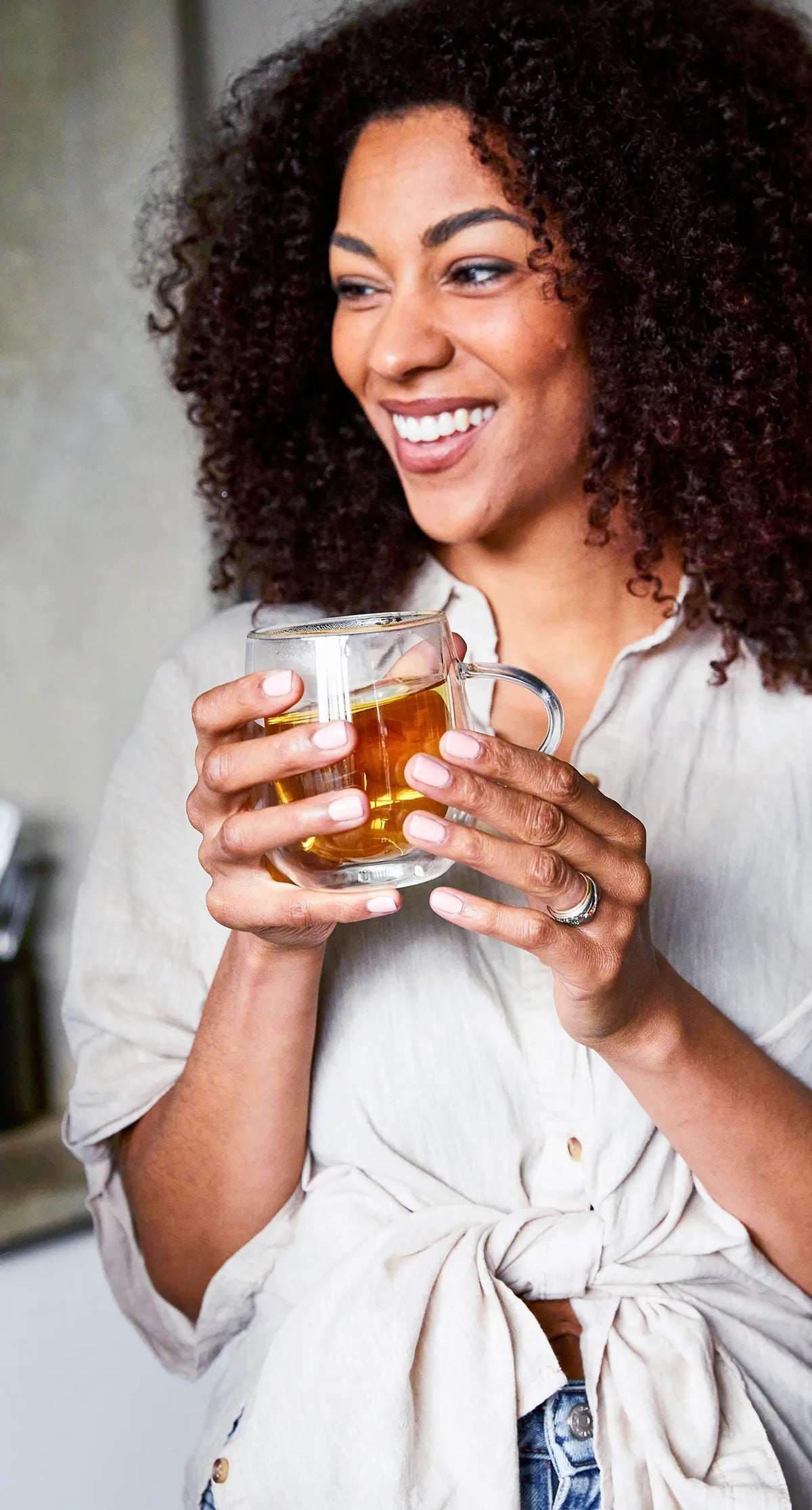 A woman with curly hair smiles while holding a clear glass mug filled with tea, wearing a light-colored shirt.
