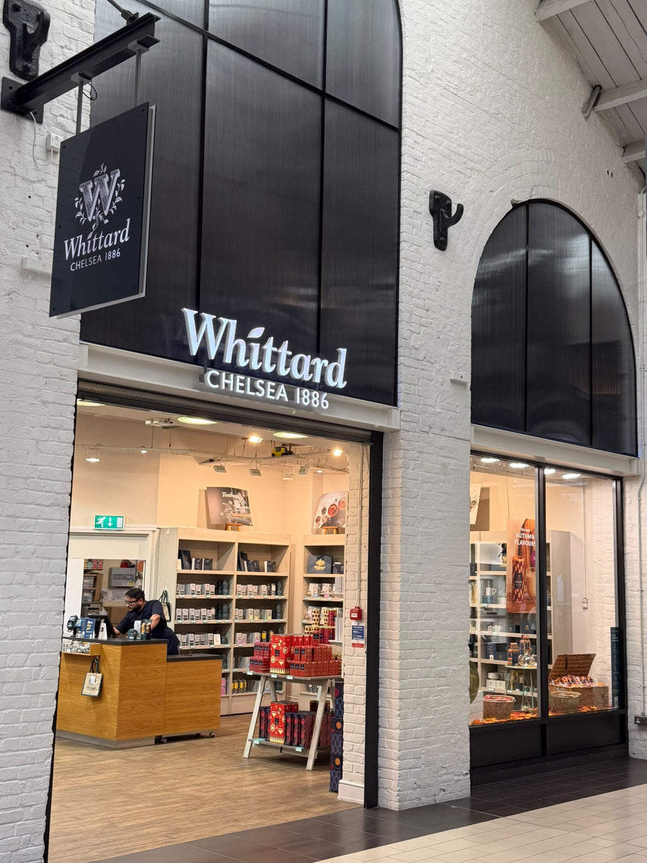 A Whittard of Chelsea 1886 store with a white brick facade, large arched windows, and a person standing behind the wooden checkout counter. Shelves display teas and related products inside.