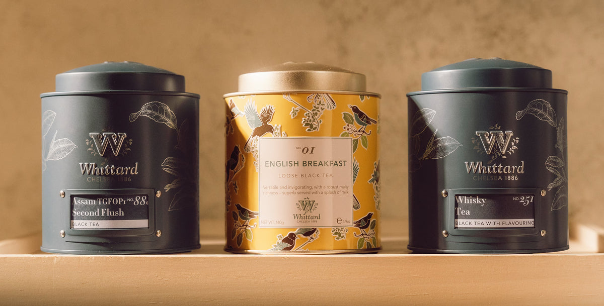 Three Whittard Chelsea 1886 tea tins on a wooden shelf: two dark grey tins labeled Assam and Whisky Tea flanking a yellow tin labeled English Breakfast.