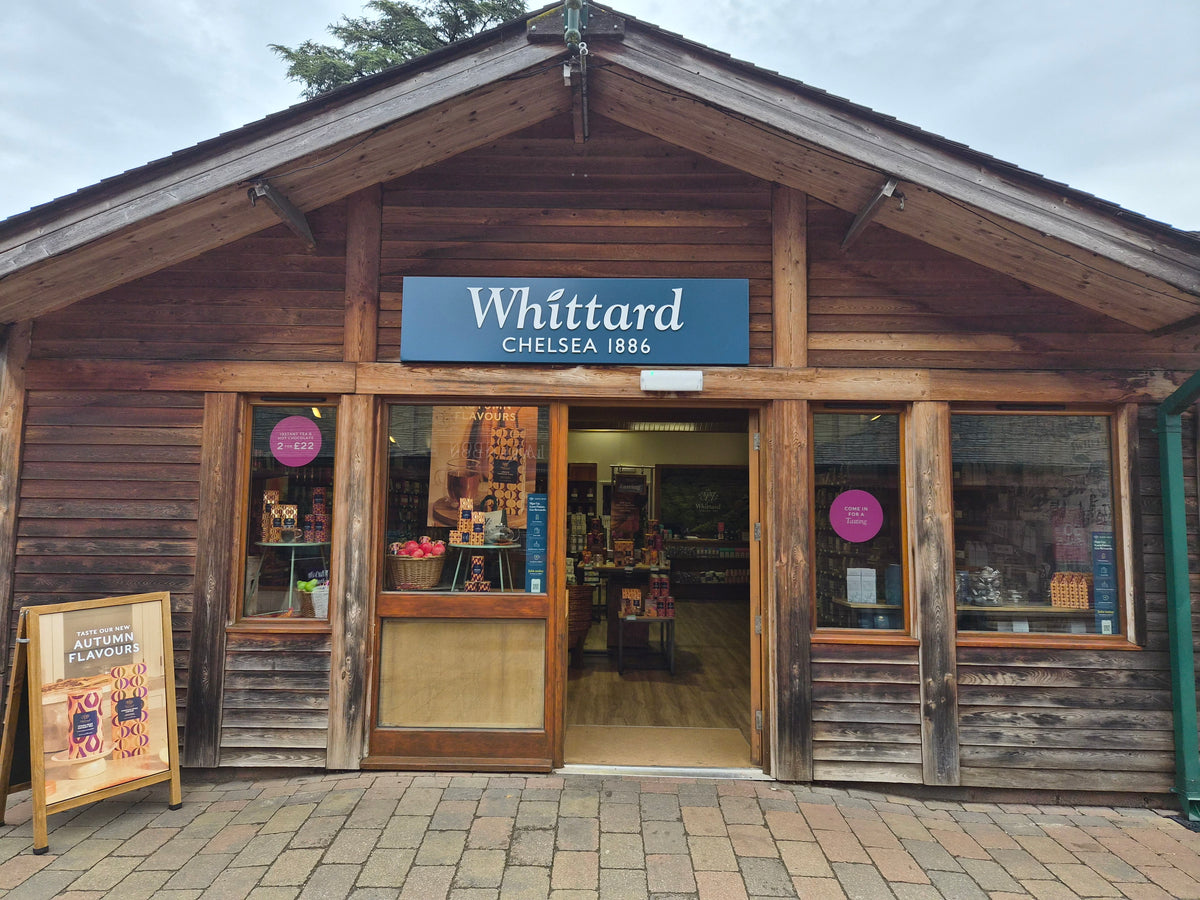 A wooden storefront with a sign reading Whittard Chelsea 1886. The door is open, displaying shelves inside. A board outside advertises autumn flavors. The exterior is rustic with two large windows.