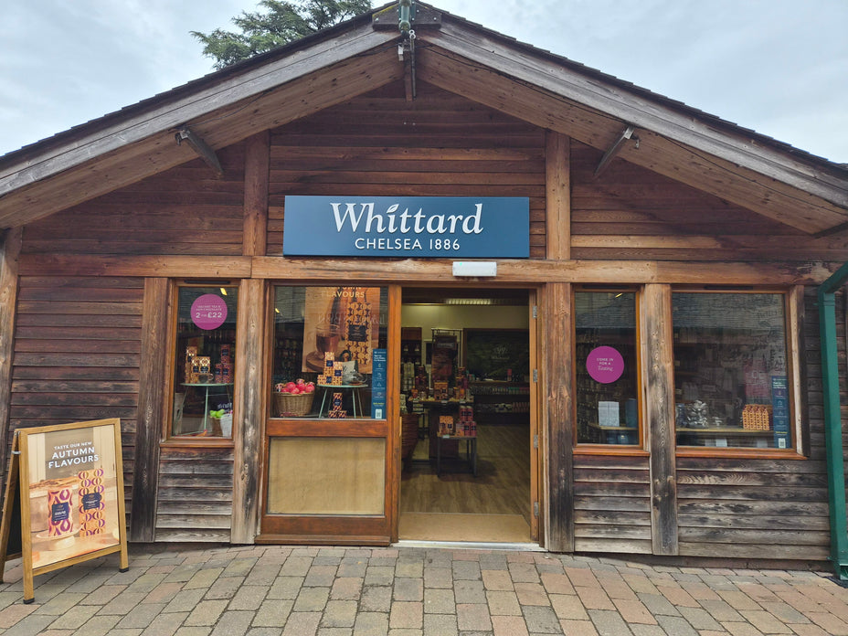 A wooden storefront with a sign reading Whittard Chelsea 1886. The door is open, displaying shelves inside. A board outside advertises autumn flavors. The exterior is rustic with two large windows.