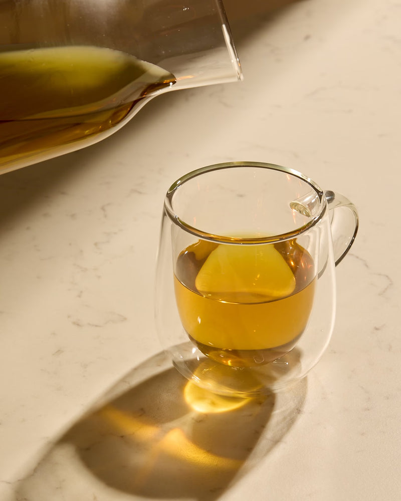 A double-walled glass cup filled with light amber liquid, sitting on a countertop as more is poured from a glass carafe above.