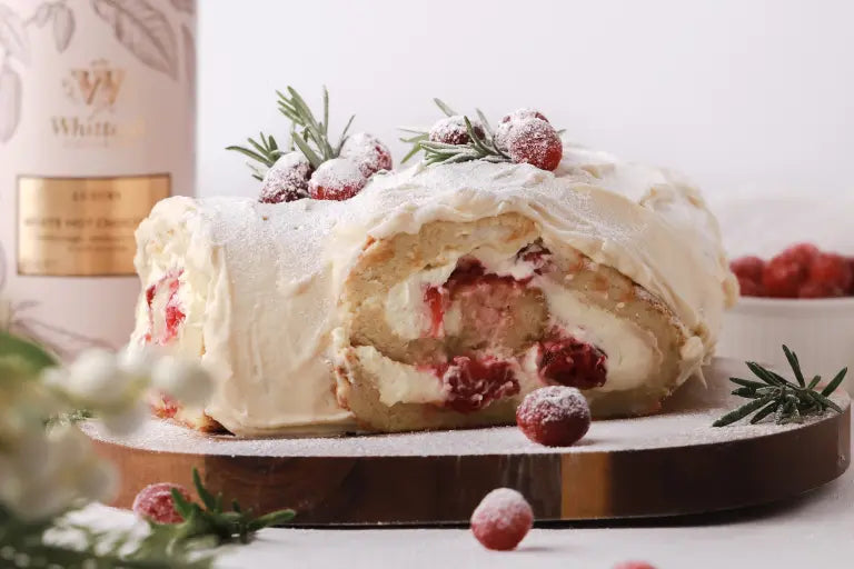 A sliced cranberry and cream roll cake topped with whipped cream, sugared cranberries, rosemary, and powdered sugar sits on a wooden board. Some cranberries and rosemary sprigs are scattered around.