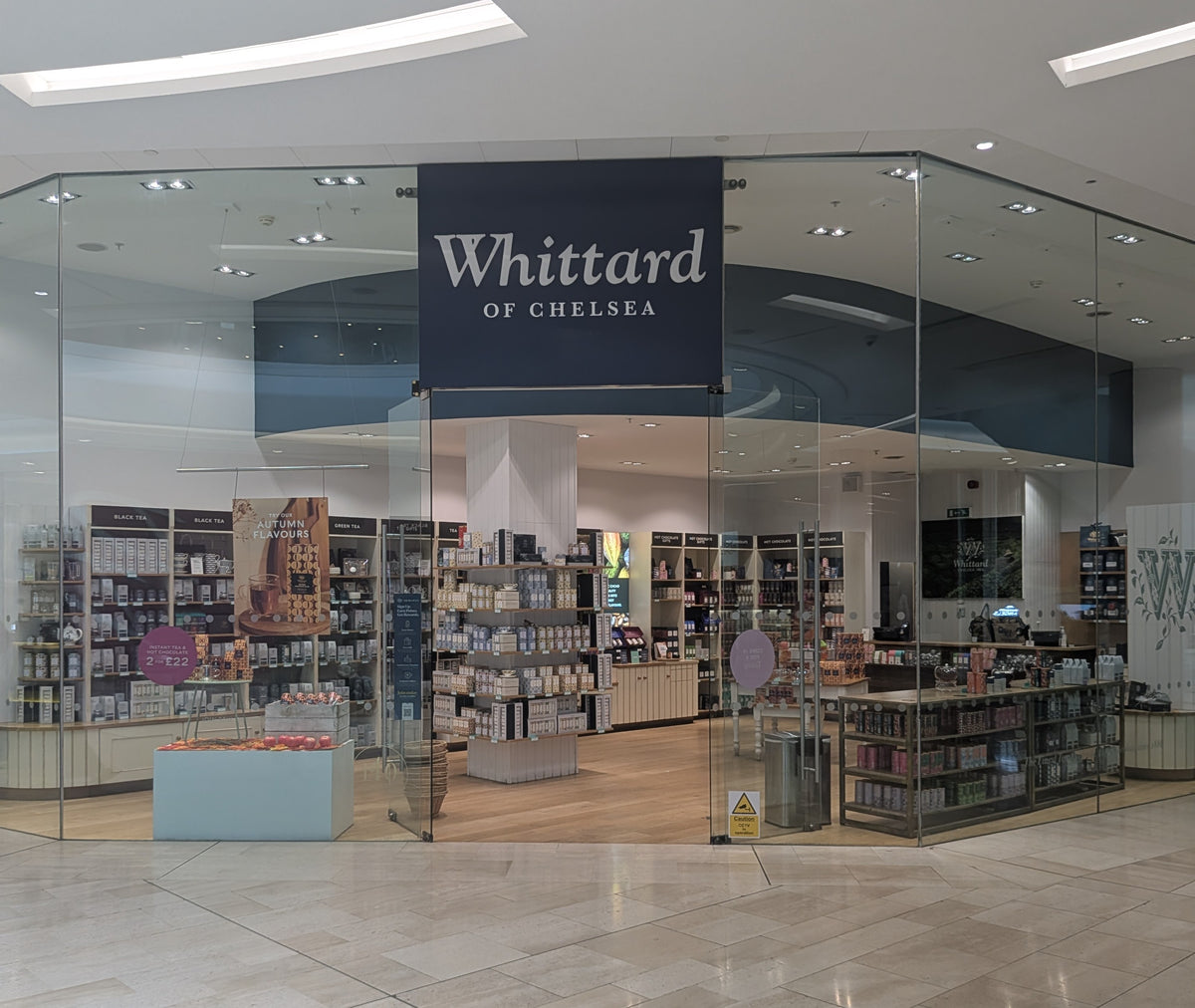A Whittard of Chelsea store with glass walls, displaying shelves filled with tea, coffee, and related products. The store has bright lighting and a modern interior design.