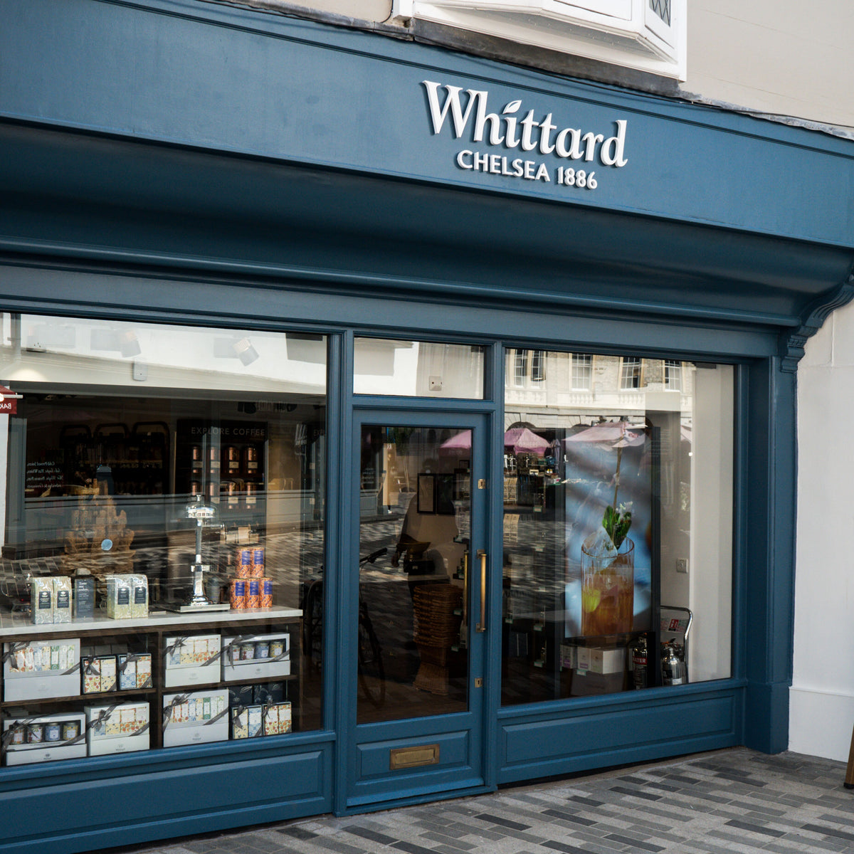 A storefront with blue trim and large glass windows displays the sign Whittard Chelsea 1886. Tea products and gift sets are visible through the window. The reflection of the street can be seen in the glass.