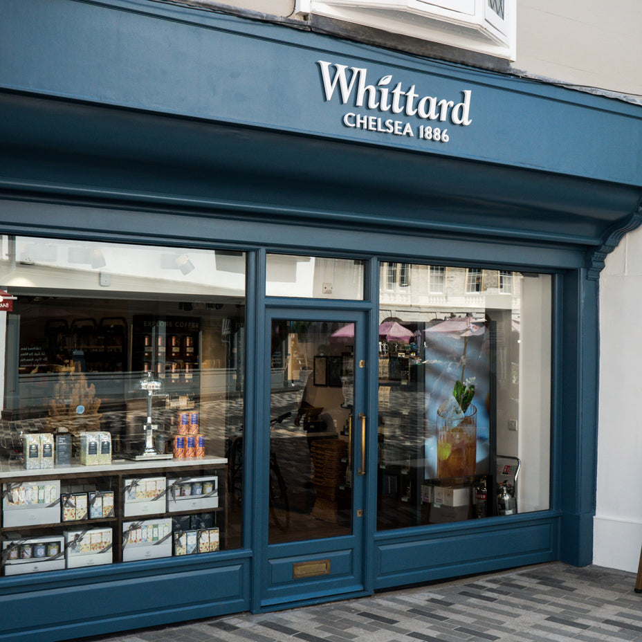 A storefront with blue trim and large glass windows displays the sign Whittard Chelsea 1886. Tea products and gift sets are visible through the window. The reflection of the street can be seen in the glass.