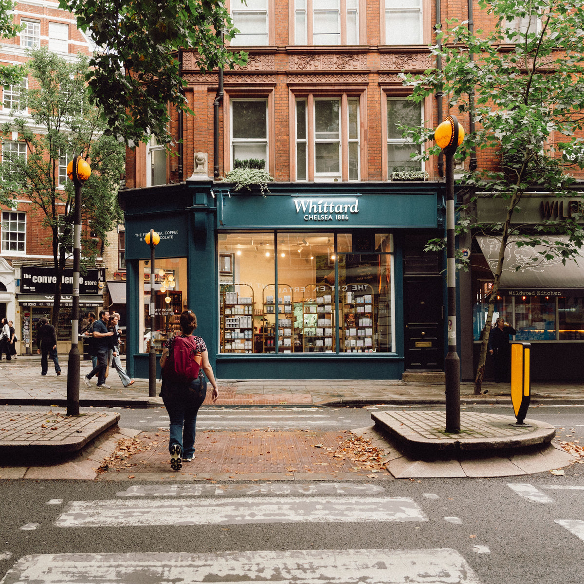 A person crosses a zebra crossing toward a blue shop called Whittard Chelsea 1886 on a city street lined with trees and brick buildings.