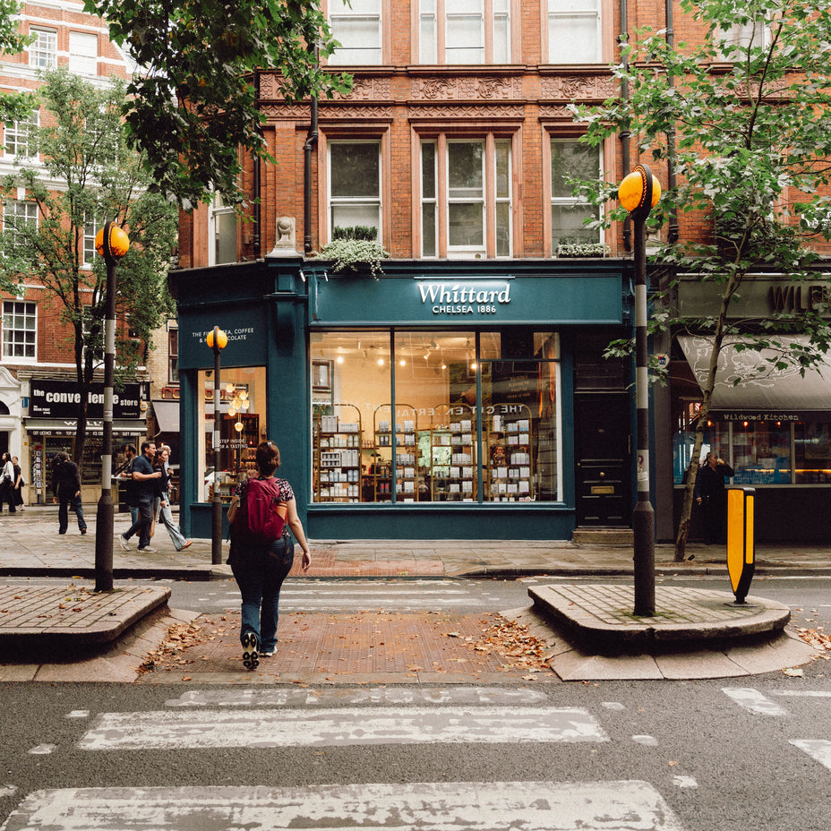 A person crosses a zebra crossing toward a blue shop called Whittard Chelsea 1886 on a city street lined with trees and brick buildings.