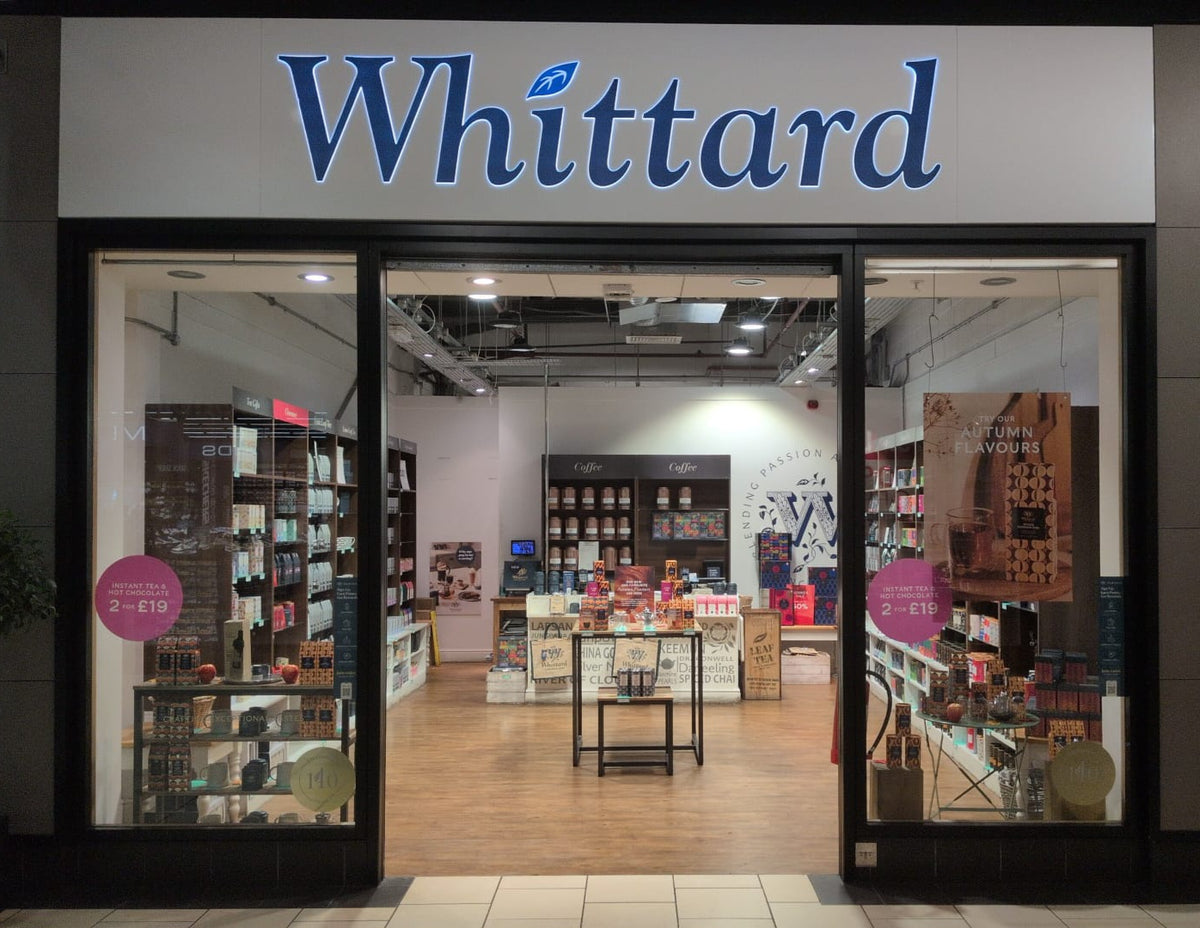A Whittard store with glass doors, tea and gift boxes on shelves, colorful displays, and bright lighting inside. Promotional signs are visible in the windows.