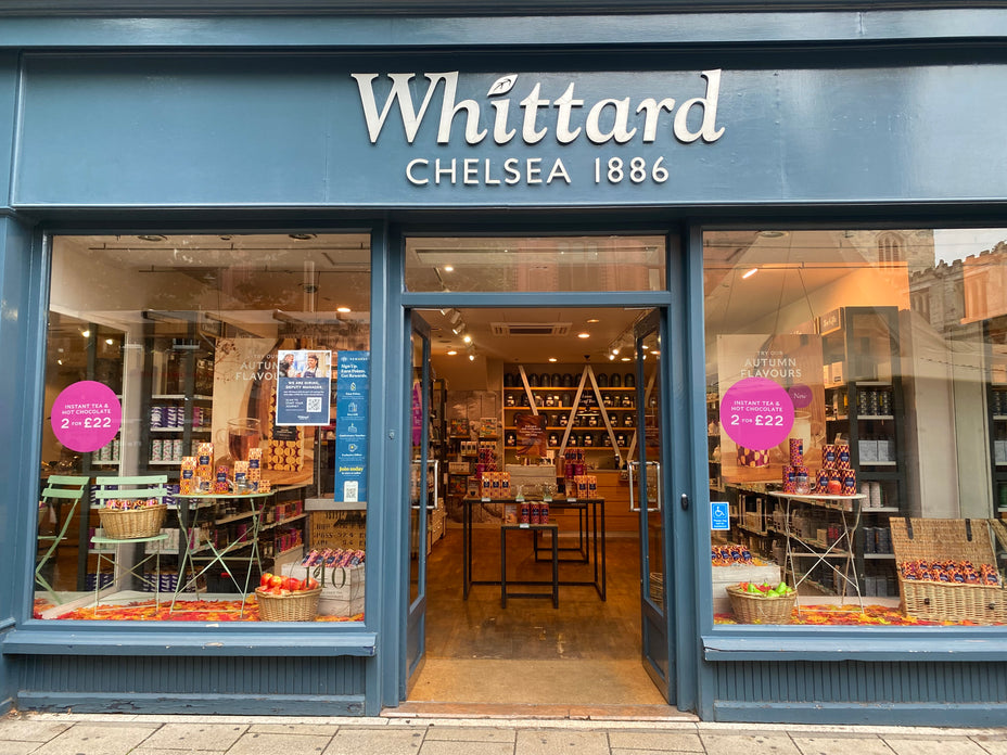 Storefront of Whittard Chelsea 1886 with glass doors, window displays of tea products and gift sets, and promotional signs reading 2 for £22. The store interior shows shelves filled with products and warm lighting.