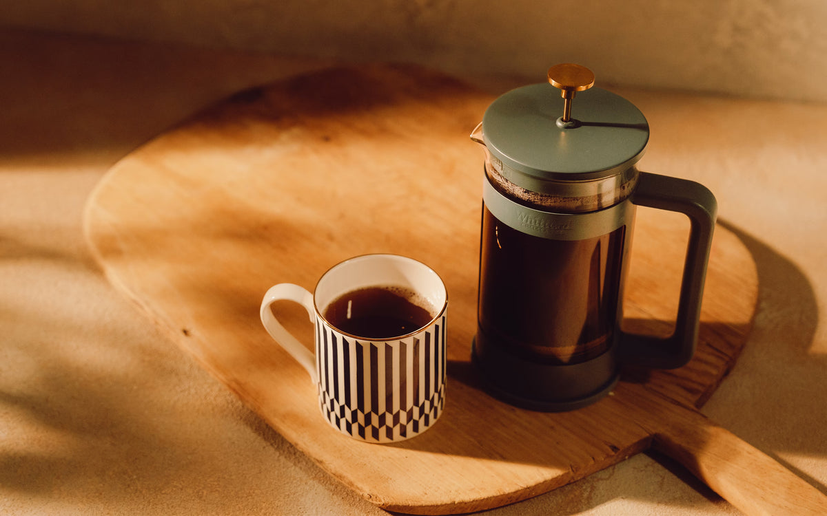 A striped mug filled with coffee next to a French press on a wooden cutting board.