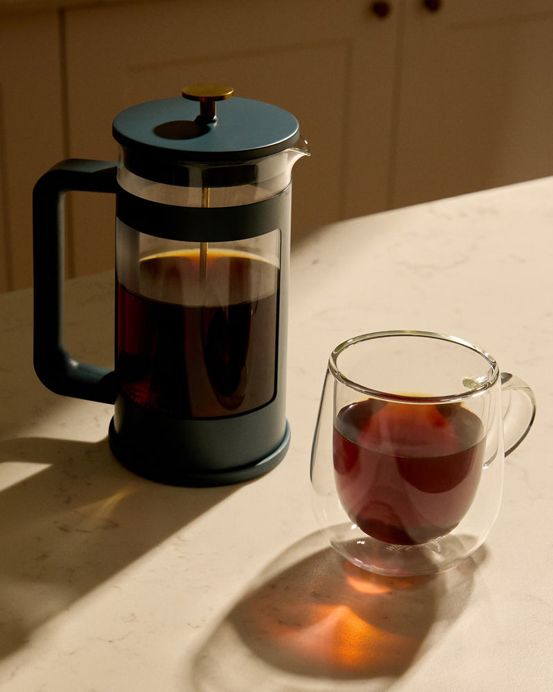 A dark blue French press next to a glass mug of brewed coffee on a light-colored countertop.