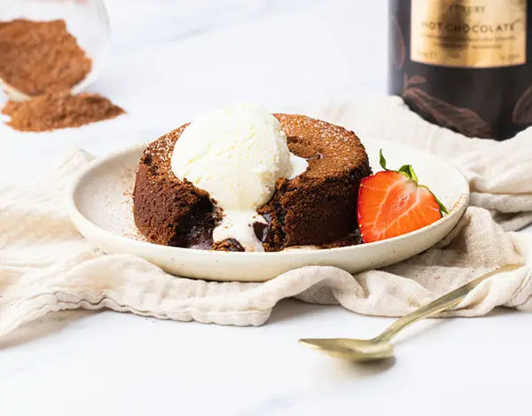 Chocolate lava cake topped with melting vanilla ice cream, served with a halved strawberry on a plate, next to a gold spoon. Cocoa powder and a hot chocolate container are visible in the background.