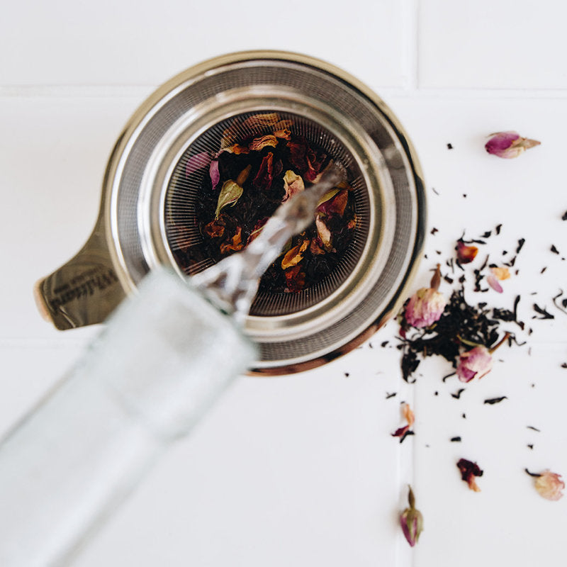 A glass bottle pours water into a metal tea infuser filled with loose tea leaves and flower petals. Some tea leaves and petals are scattered on the white surface nearby.