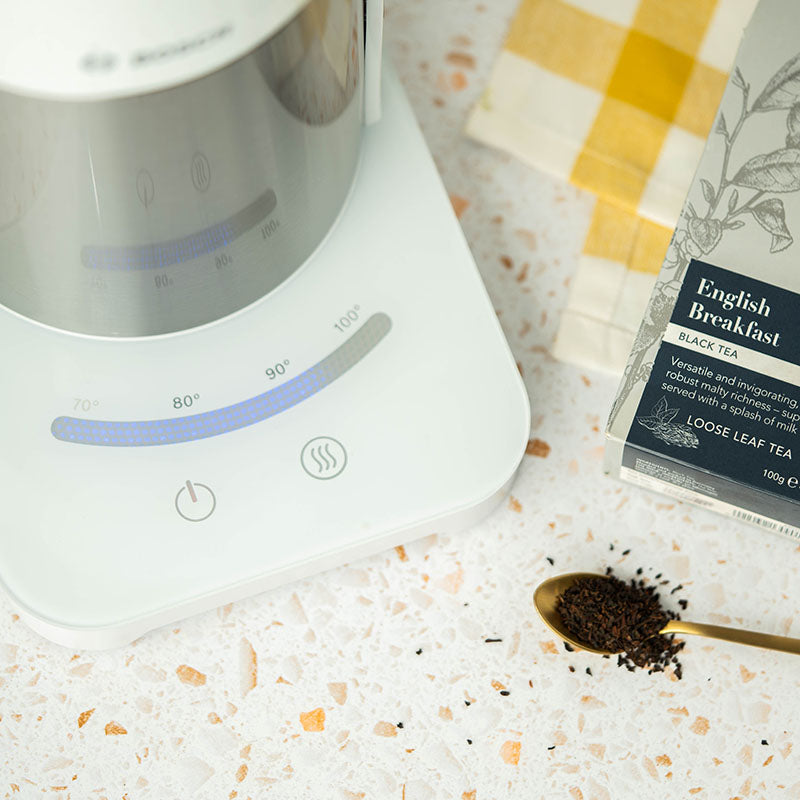 A white electric kettle sits on a terrazzo countertop near a gold spoon with loose leaf tea, a box of English Breakfast black tea, and a yellow checked napkin.