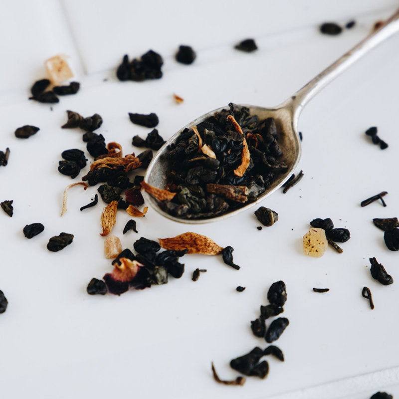 A metal spoon holding loose leaf tea with herbs and dried fruit, resting on a white surface scattered with more tea leaves and ingredients.