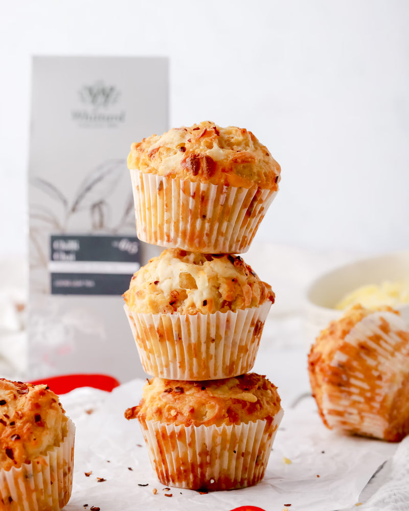 Three cheese muffins stacked on top of each other on parchment paper, with another muffin lying on its side and a box in the background.
