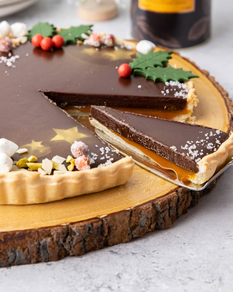 A chocolate tart with a caramel layer, decorated with holly leaves, red berries, marshmallows, and nuts, sits on a wooden board. A slice is being lifted from the tart, revealing the layers inside.