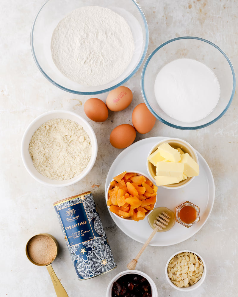 A flat lay of baking ingredients: bowls of flour, sugar, ground almonds, dried apricots, eggs, butter, honey, a canister of tea, light brown sugar, slivered almonds, and dried cranberries arranged on a white surface.