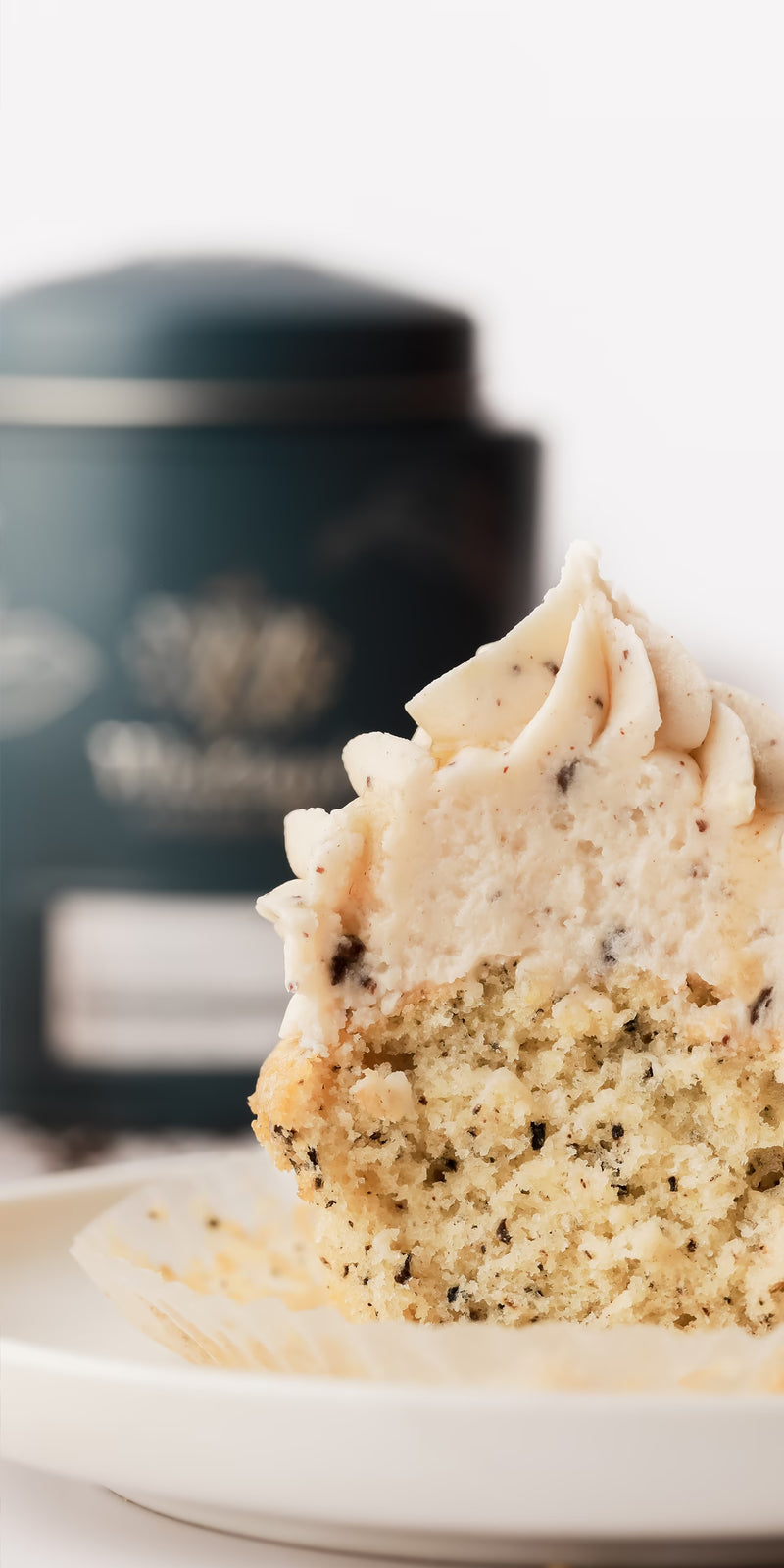 A close-up of a cupcake with swirled frosting and visible poppy seeds, partially unwrapped on a plate, with a blurred dark container in the background.