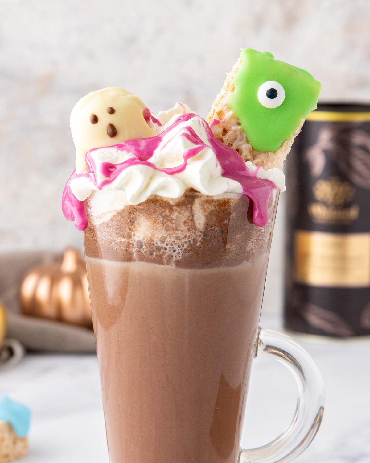 A glass mug of hot chocolate topped with whipped cream, pink drizzle, and two decorated treats: one shaped like a ghost and one like a green monster with one eye.