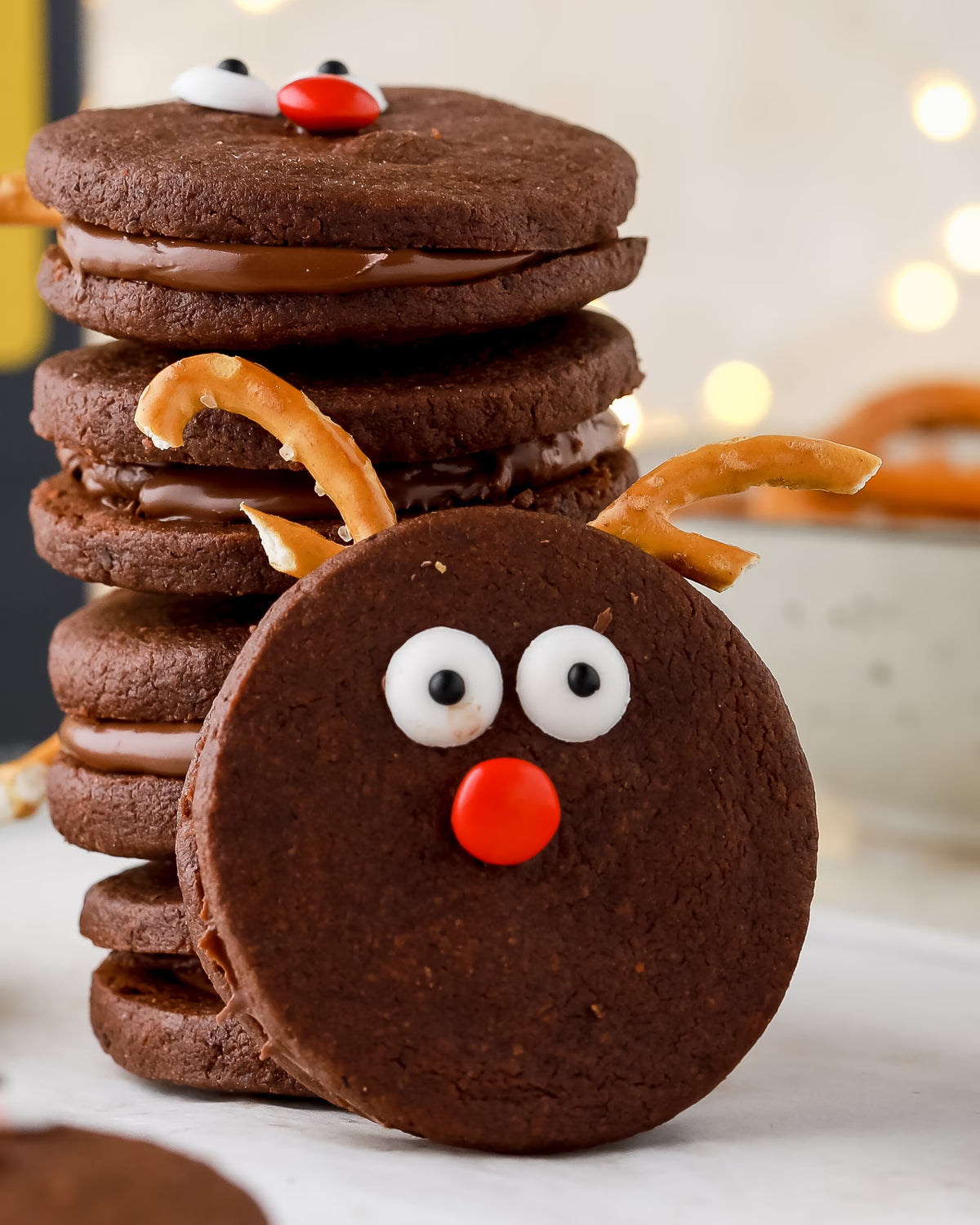 Chocolate sandwich cookies decorated as reindeer with candy eyes, red candy noses, and pretzel antlers are stacked on a white surface with blurred lights in the background.