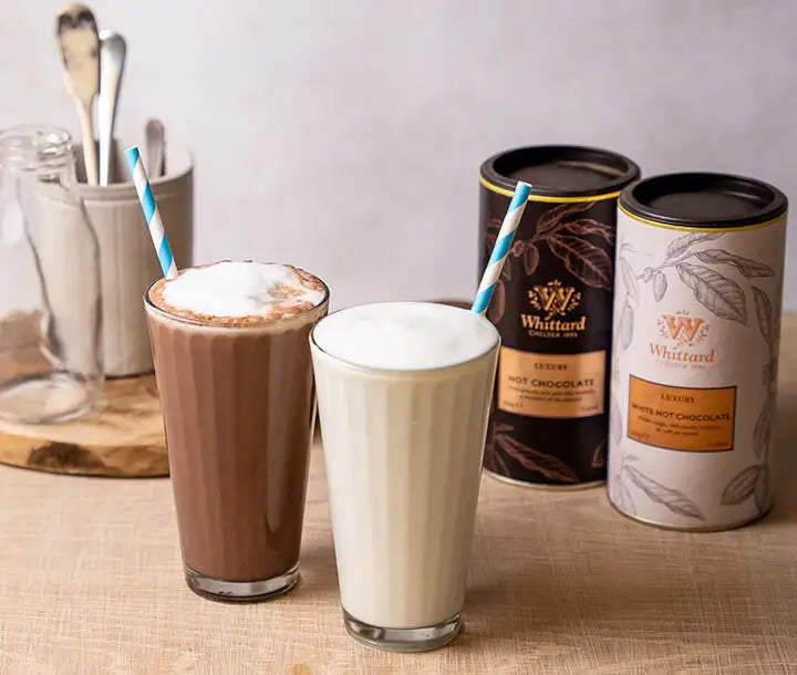 Two glasses with blue-striped straws contain chocolate and white hot chocolate drinks. Behind them are two Whittard tins, one brown and one beige, placed on a light surface next to a jar holding spoons.