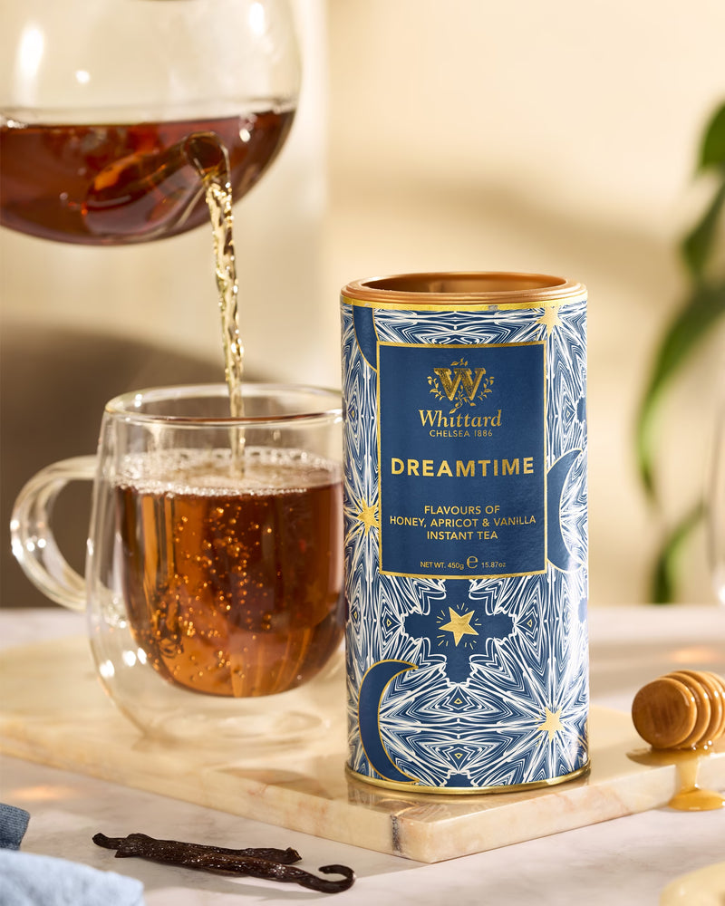 Tea being poured from a glass pot into a double-walled mug next to a tin of Whittard Dreamtime instant tea.