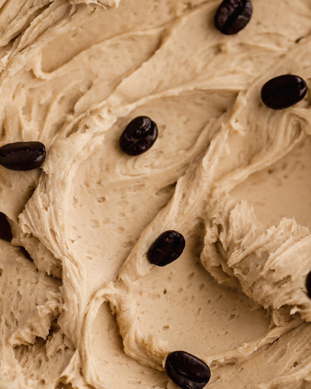 Close-up of creamy, light brown coffee-flavored frosting or mousse, textured with swirls and decorated with whole coffee beans scattered on top.