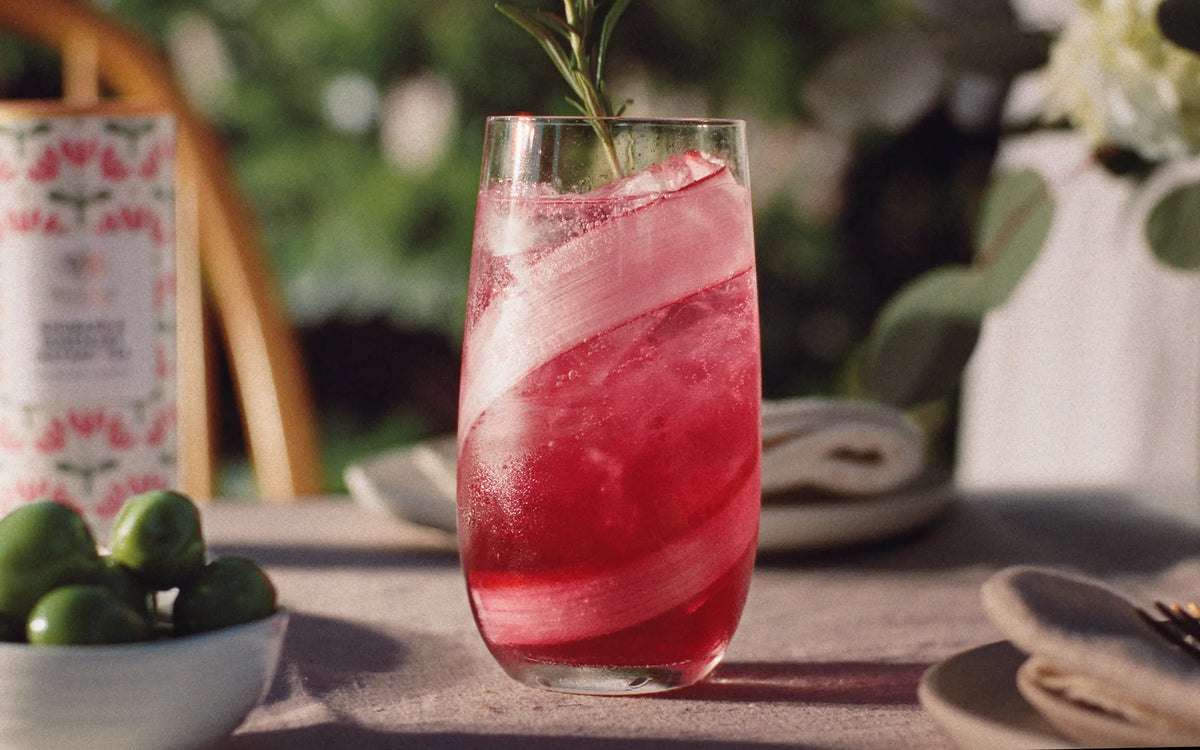 A tall glass of pink sparkling drink with a large ice ribbon and a rosemary sprig sits on a table, surrounded by blurred dishes and greenery in the background.