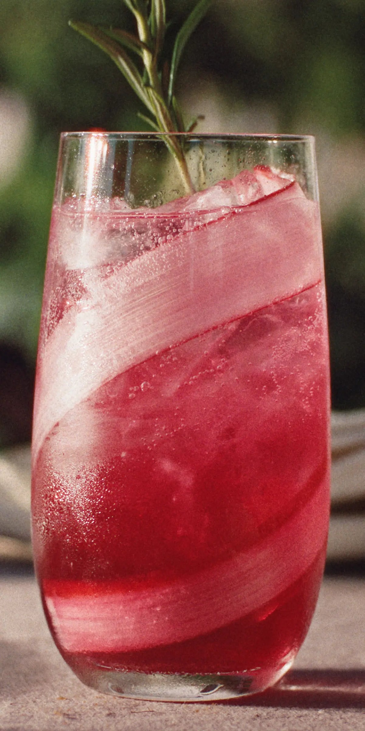 A glass of red sparkling drink with ice, garnished with a rosemary sprig. The glass has a frosty swirl design and is set on a surface with a blurred green background.
