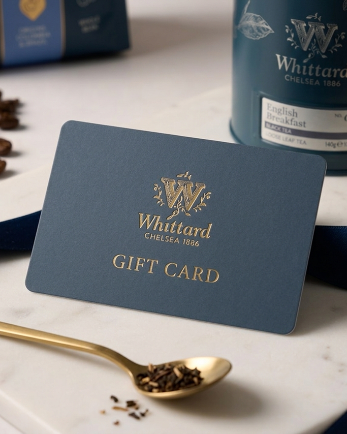 A Whittard E-Gift Card in blue sits elegantly on a white surface beside a gold spoon holding loose tea leaves, with a blue tin of English Breakfast tea set in the background—making it the ideal gift for any tea lover. You can even include a personal message for an extra special touch.