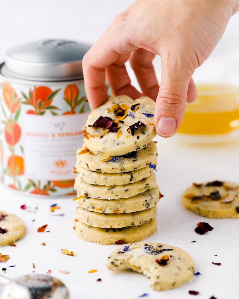 A hand picks up a floral shortbread cookie from a stack. Cookies are decorated with dried flower petals. A tin labeled Mango & Bergamot and a cup of tea sit in the background.