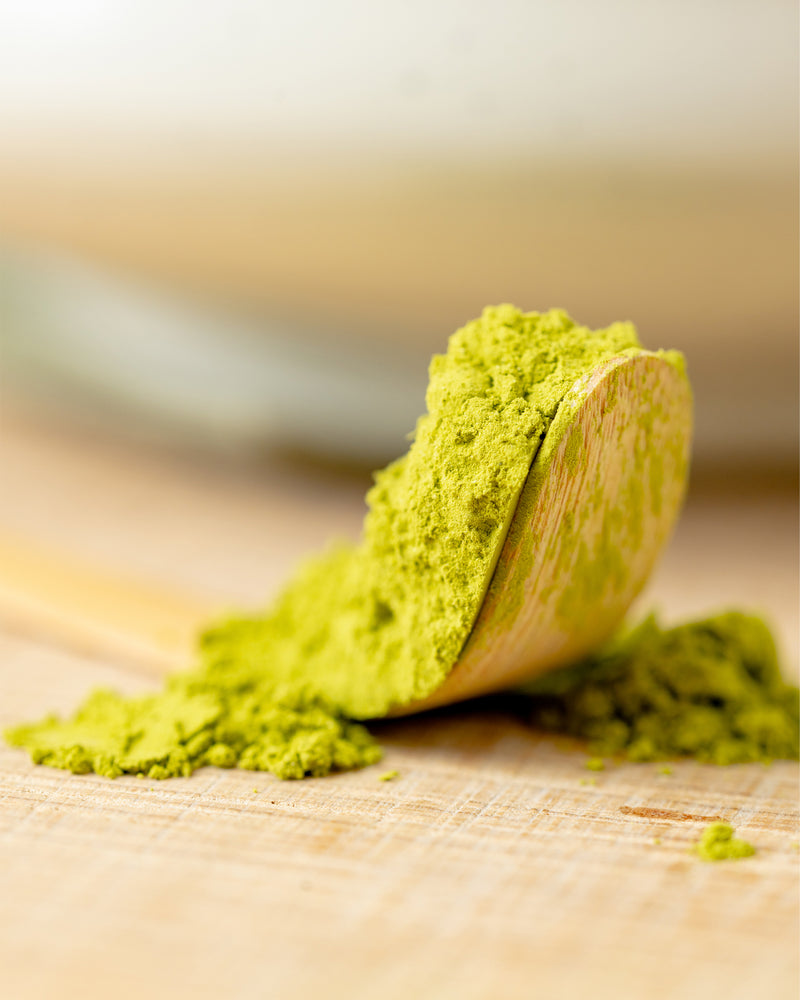 A wooden spoon rests on a light wooden surface, filled with vibrant green matcha powder, with some powder spilling onto the surface.