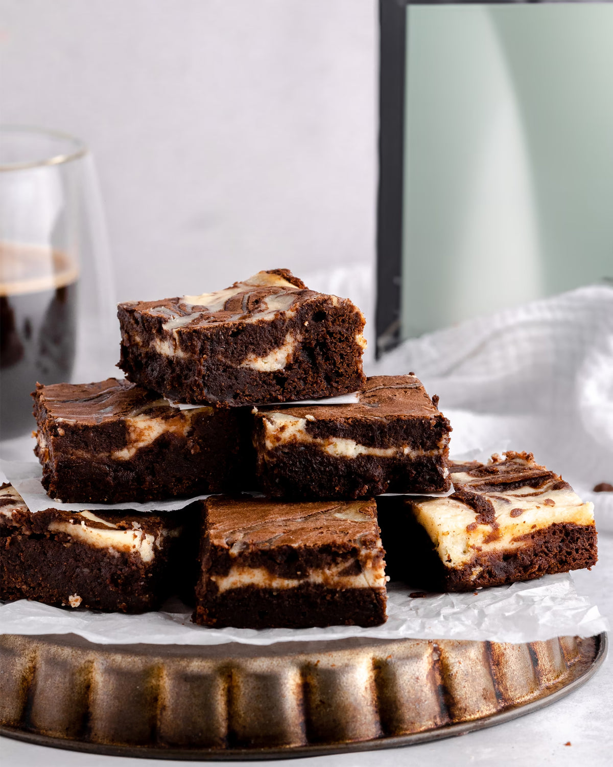 A stack of chocolate brownies with a cream cheese swirl sits on a round tray lined with parchment paper. A glass of dark drink and a mirror are visible in the background.