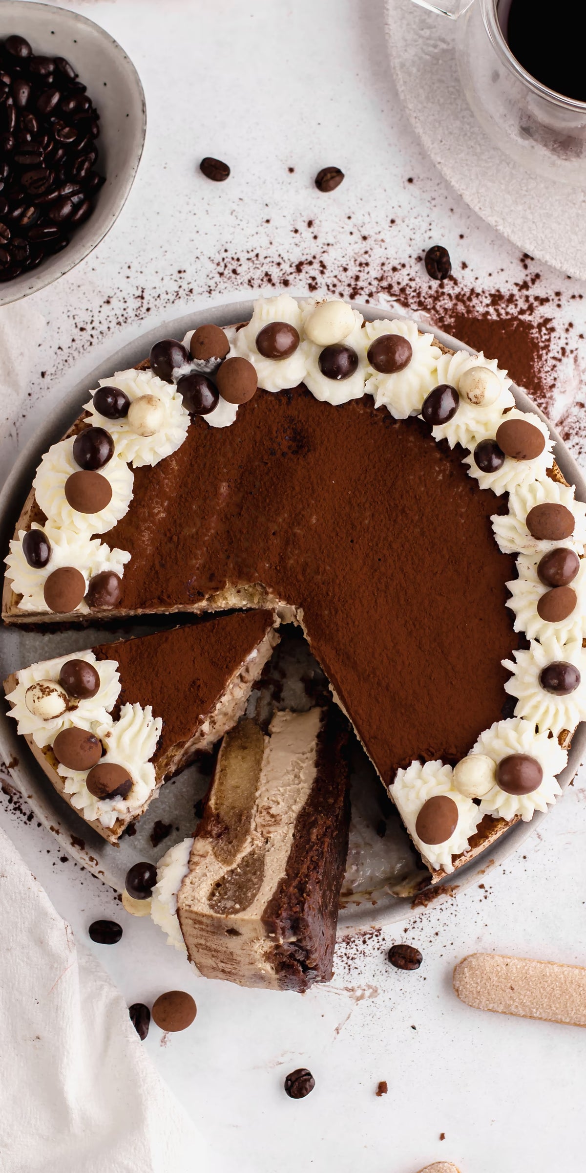 A round tiramisu cake topped with whipped cream, chocolate balls, and cocoa powder. A slice is cut and lifted slightly from the cake, showing creamy and layered insides. Coffee beans and cocoa powder are scattered nearby.