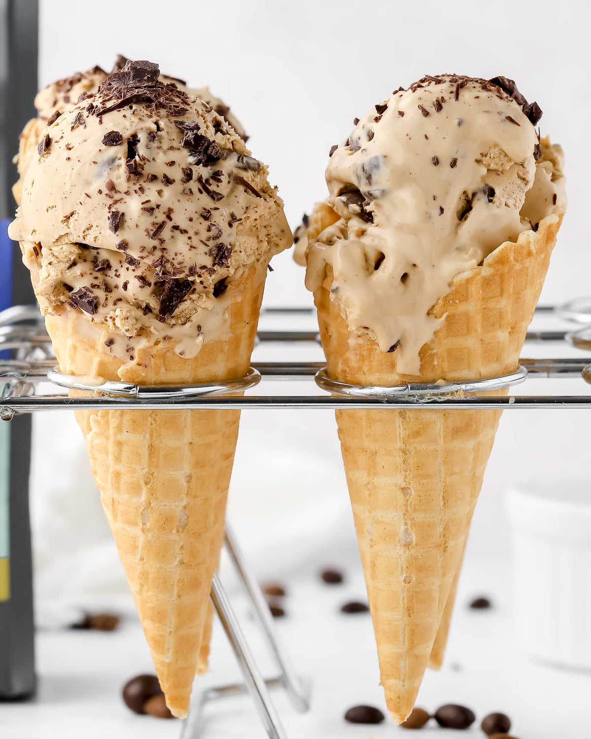 Two waffle cones filled with creamy coffee ice cream, topped with chocolate shavings, are displayed in a metal holder with some coffee beans scattered in the background.