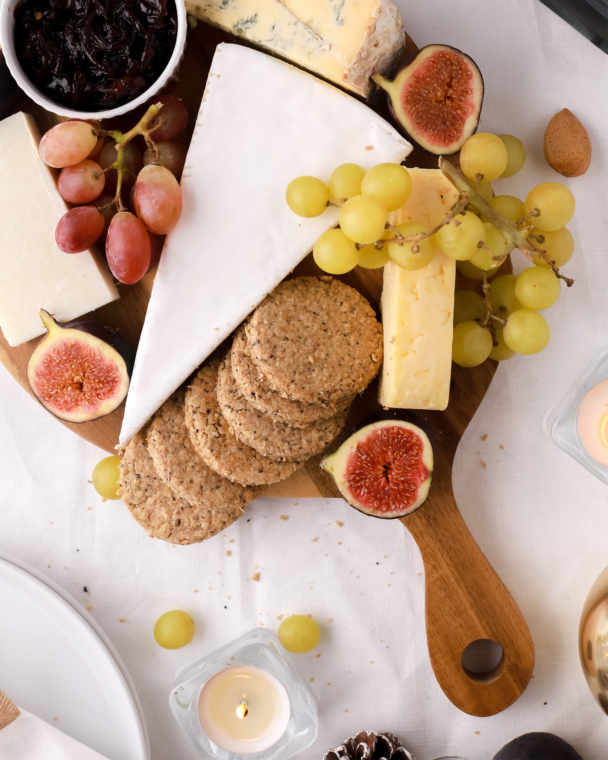 A cheese board with various cheeses, grapes, figs, crackers, a bowl of dried fruit, and lit candles on a white tablecloth.