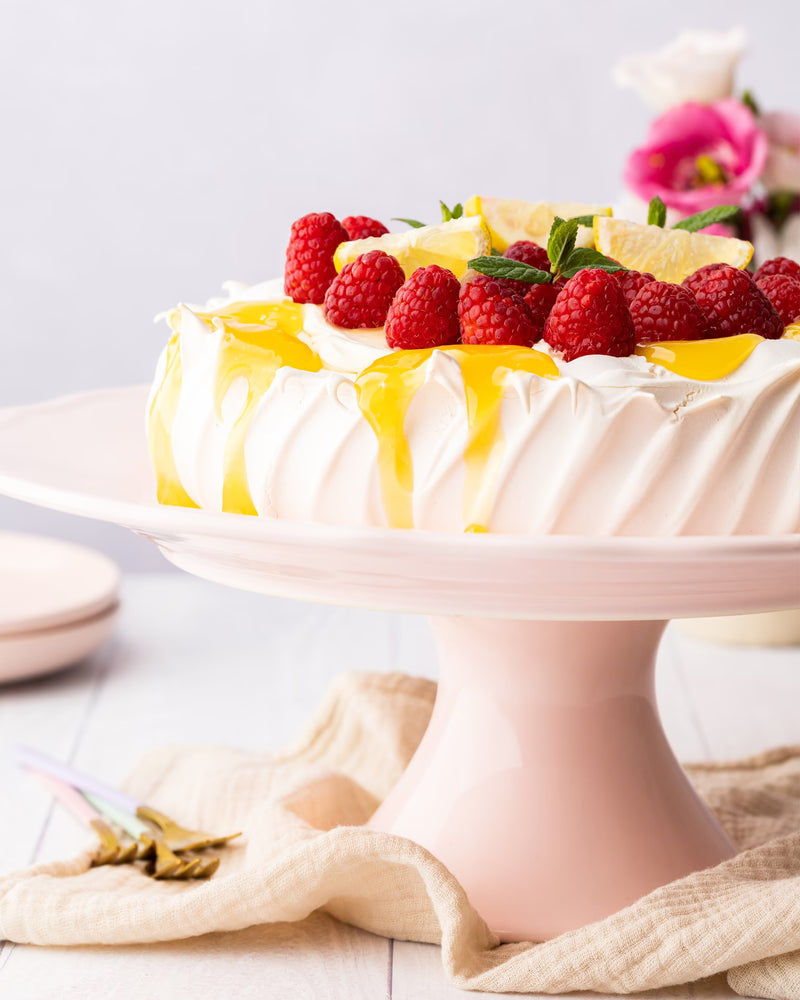 A pavlova topped with raspberries, lemon slices, mint, and yellow sauce sits on a pale pink cake stand, with a beige cloth and flowers in the background.