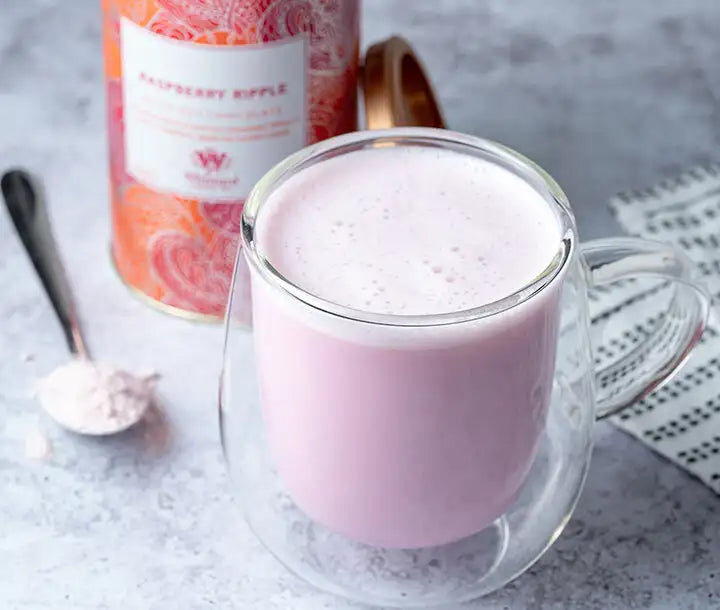 A clear glass mug filled with pink raspberry-flavored latte sits on a gray surface. Next to it is a tin labeled Raspberry Ripple, a spoon with pink powder, and a white cloth with black stripes.