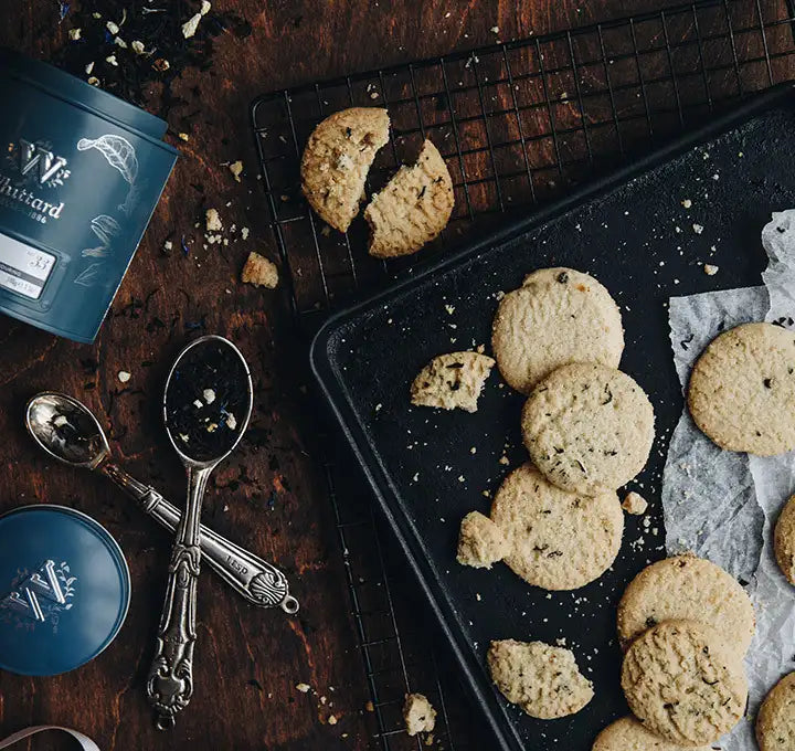 Chocolate chip cookies on a baking tray and cooling rack, some broken, with cookie crumbs scattered. Nearby are vintage spoons and a blue container on a rustic wooden table.