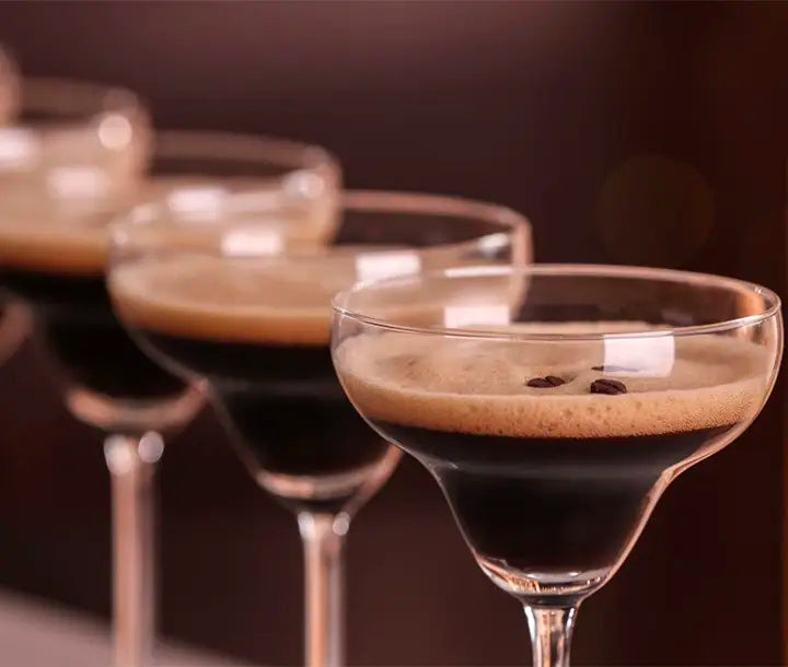 Four coupe glasses filled with dark espresso martinis are lined up in a row, each cocktail topped with a creamy foam and garnished with coffee beans. The background is softly blurred.