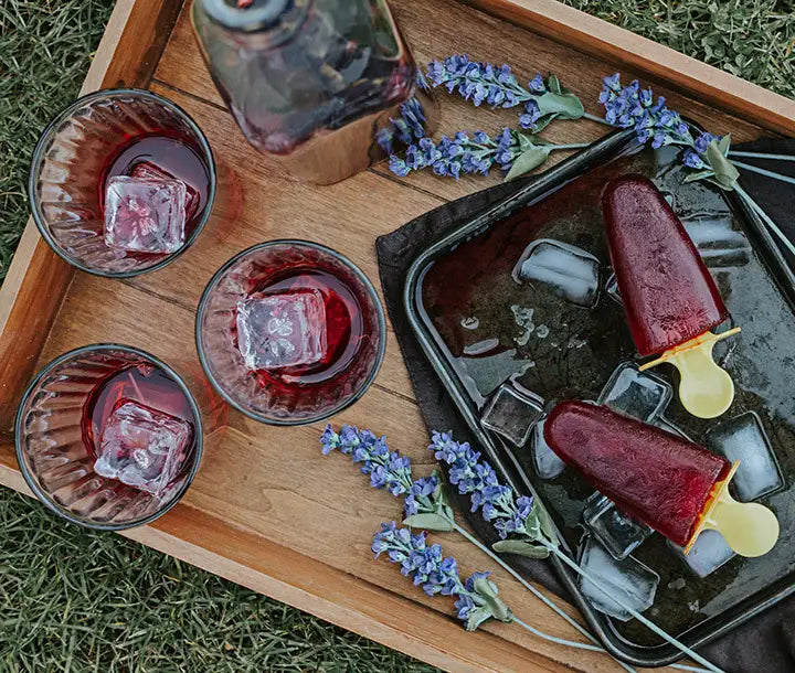 A wooden tray with two red popsicles, three glasses of iced red drink, purple flowers, and some ice cubes on a grassy surface.