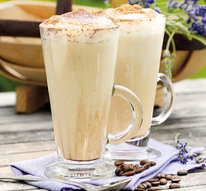 Two glass mugs filled with frothy cappuccino topped with cinnamon, placed on a napkin with coffee beans and a spoon on a wooden table, with flowers and a wooden bowl in the background.