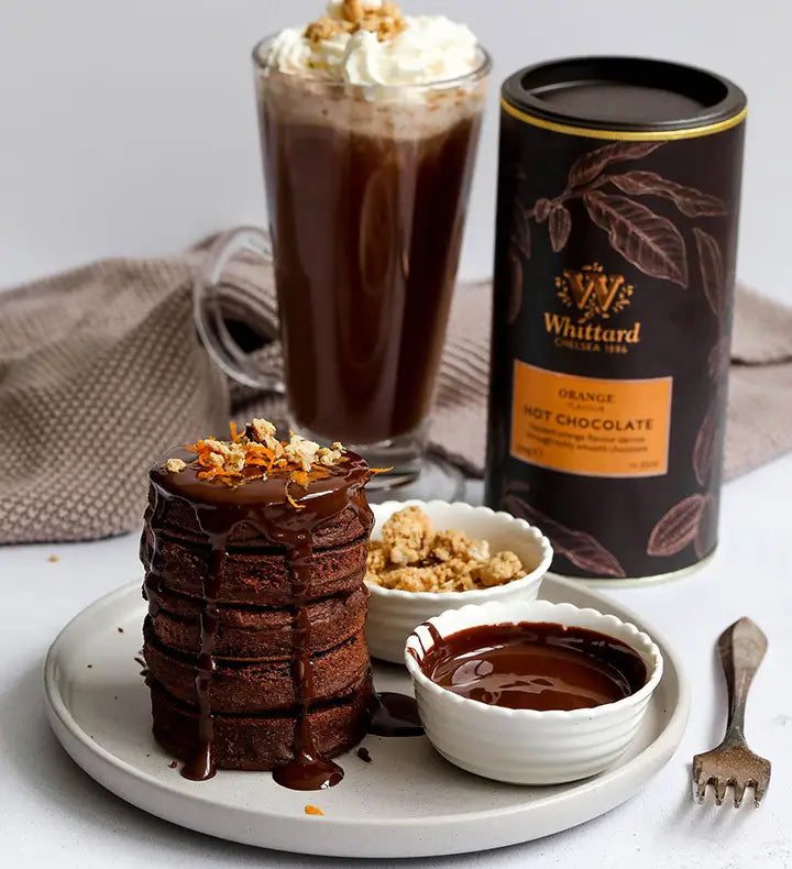 A stack of chocolate pancakes with chocolate sauce, chopped nuts, a bowl of sauce, crumbled topping, a canister of orange hot chocolate, and a glass of whipped cream-topped hot chocolate on a white plate.