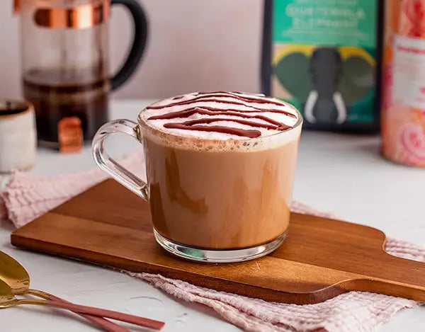 A glass mug of frothy coffee topped with chocolate drizzle sits on a wooden board, with a French press and coffee containers in the background.